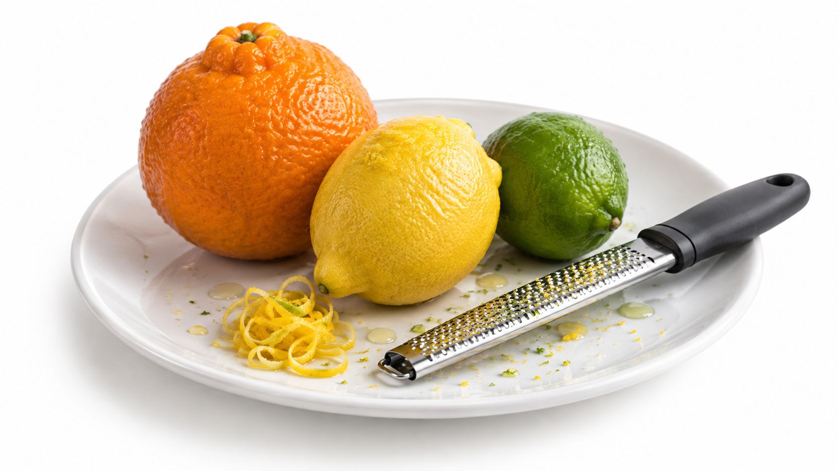 A plate featuring an orange, a lemon, a lime, and a grater with lemon zest on display.
